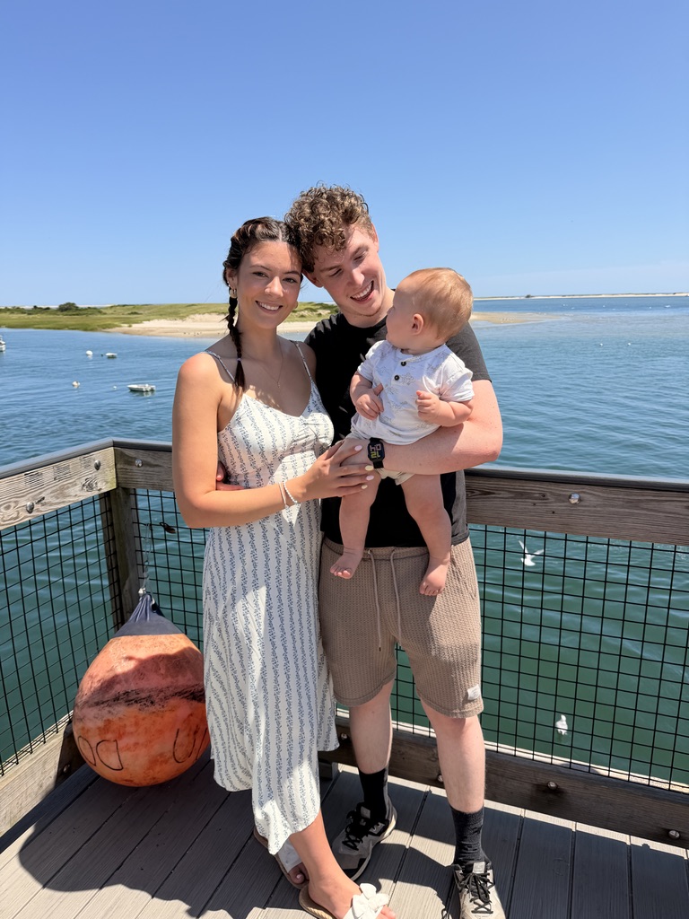 Sam standing with his wife and baby beside the water on a sunny day